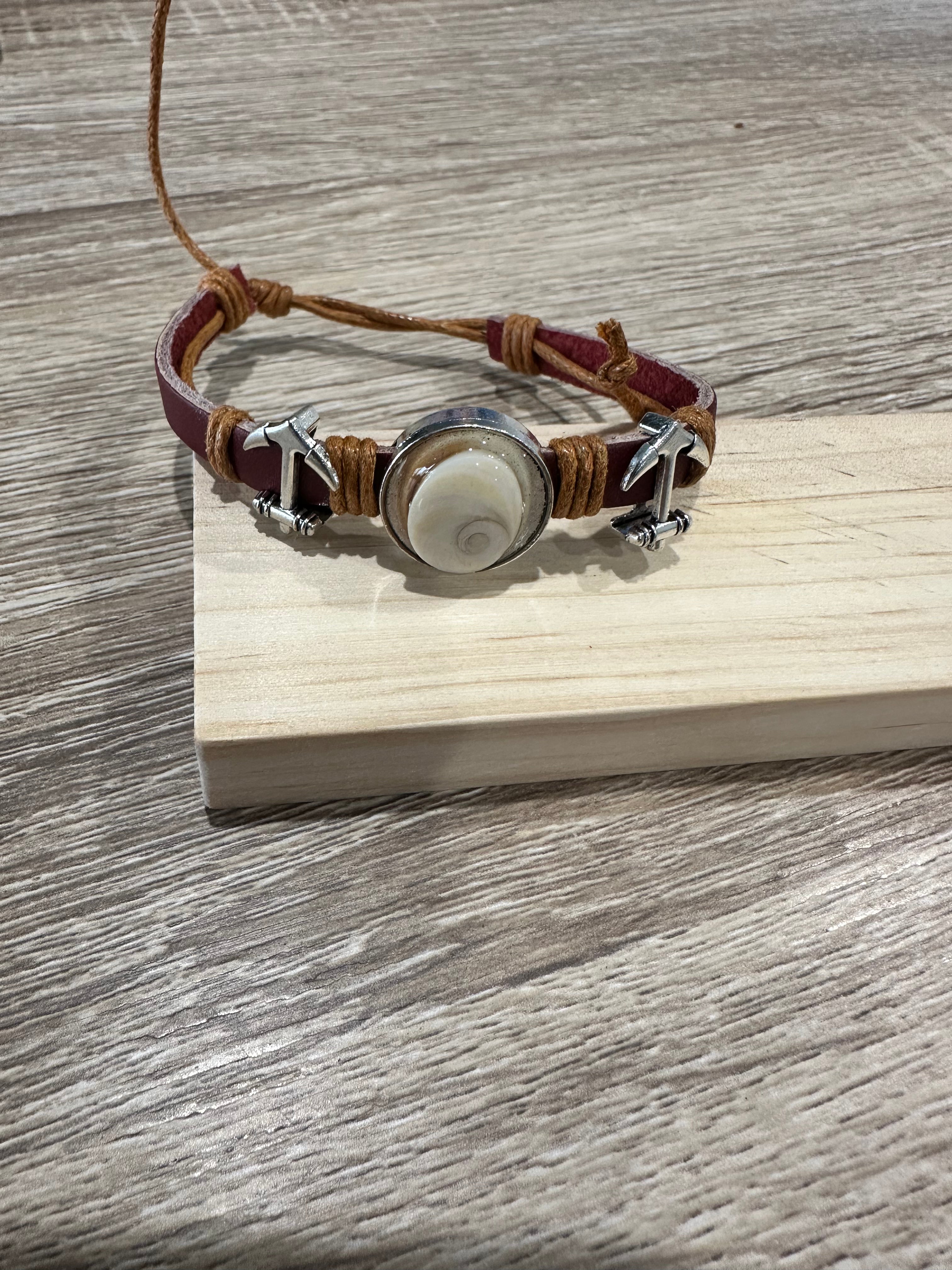 Bracelet ajustable en cuir marron et cordon beige tressé, avec deux breloques en forme d'ancre marine argentée de part et d'autre d'un cabochon rond serti en métal, contenant un œil de Sainte-Lucie (coquillage spiralé blanc). Présenté sur un support en bois clair.