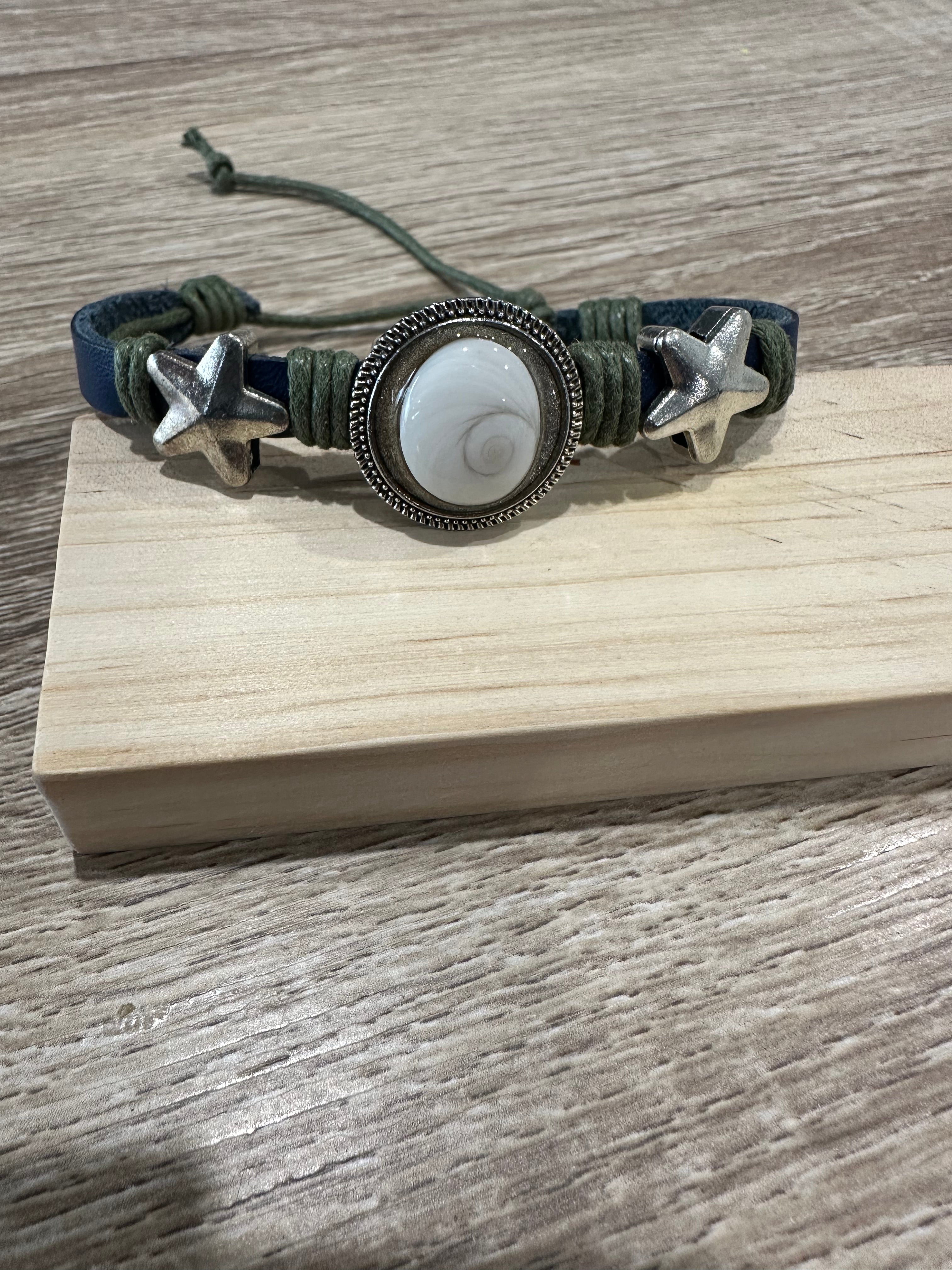 Bracelet ajustable en cuir bleu marine et cordon vert kaki tressé, avec deux breloques en forme d'étoile de mer argentée de chaque côté d'un grand cabochon rond serti en métal ciselé, contenant un œil de Sainte-Lucie (coquillage spiralé blanc nacré). Présenté sur un support en bois clair.