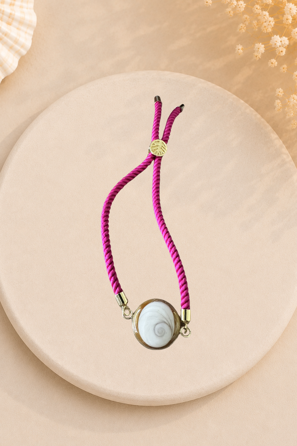 Bracelet femme cordon fuchsia au poignet, médaillon rond doré serti d’un Œil de Sainte‑Lucie blanc, embouts dorés, style chic et marin sur fond gris.