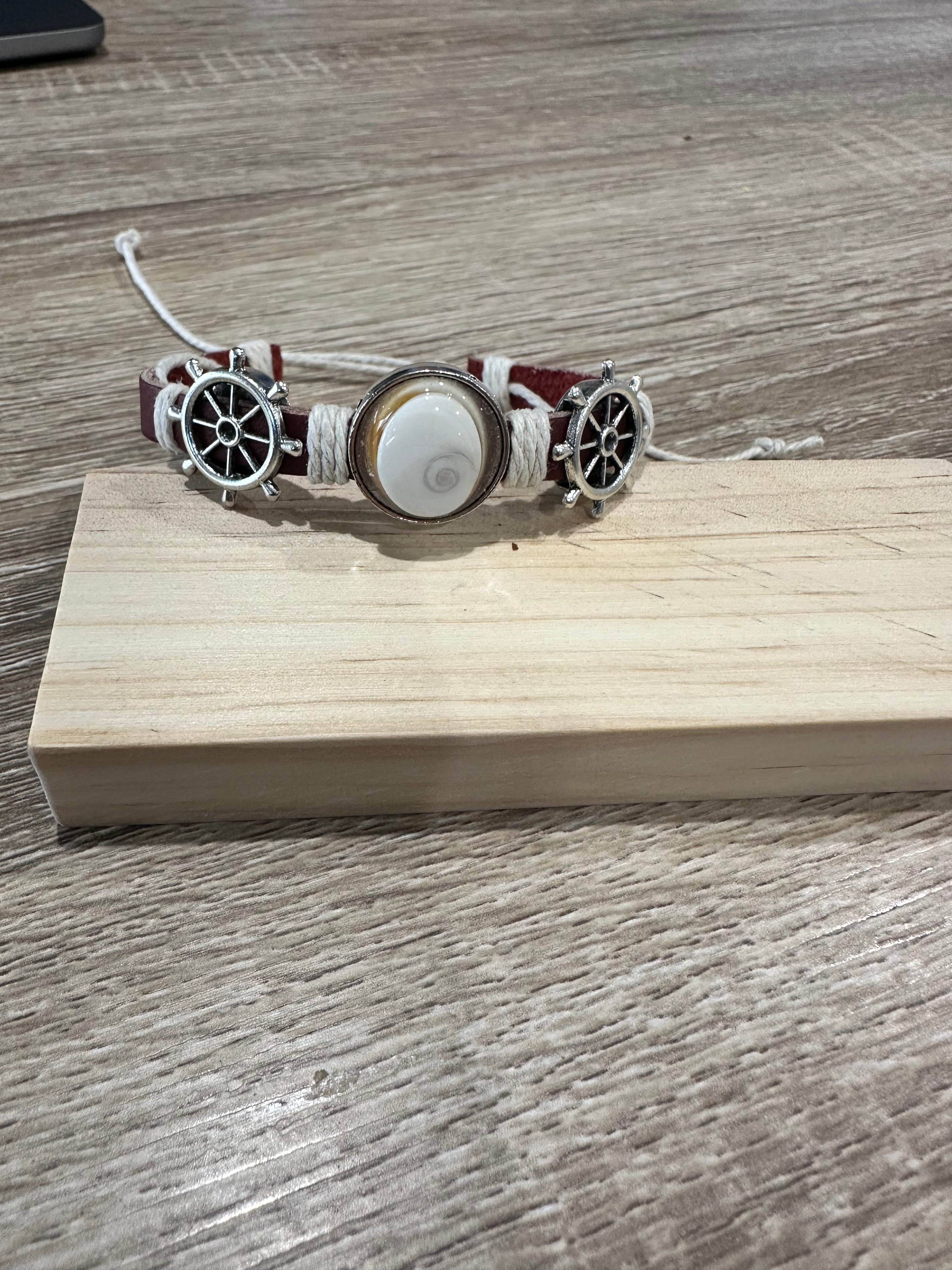 Bracelet en cuir bordeaux orné d'un Œil de Sainte-Lucie et de deux breloques barre à roue argentées, posé sur un support en bois