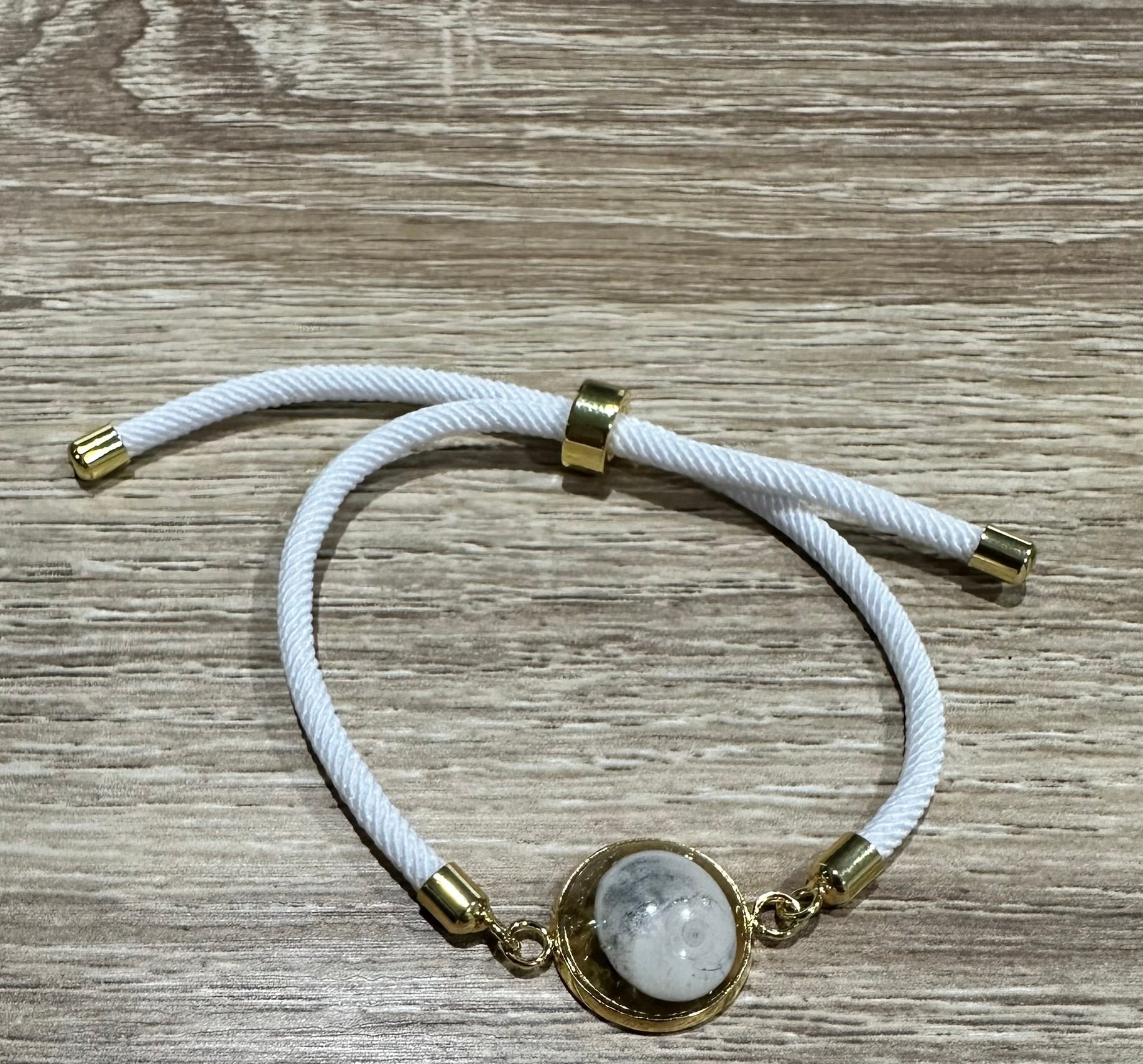 Bracelet femme sur coussin gris, cordon blanc fin, médaillon rond doré serti d’un Œil de Sainte‑Lucie blanc, style chic et minimaliste.