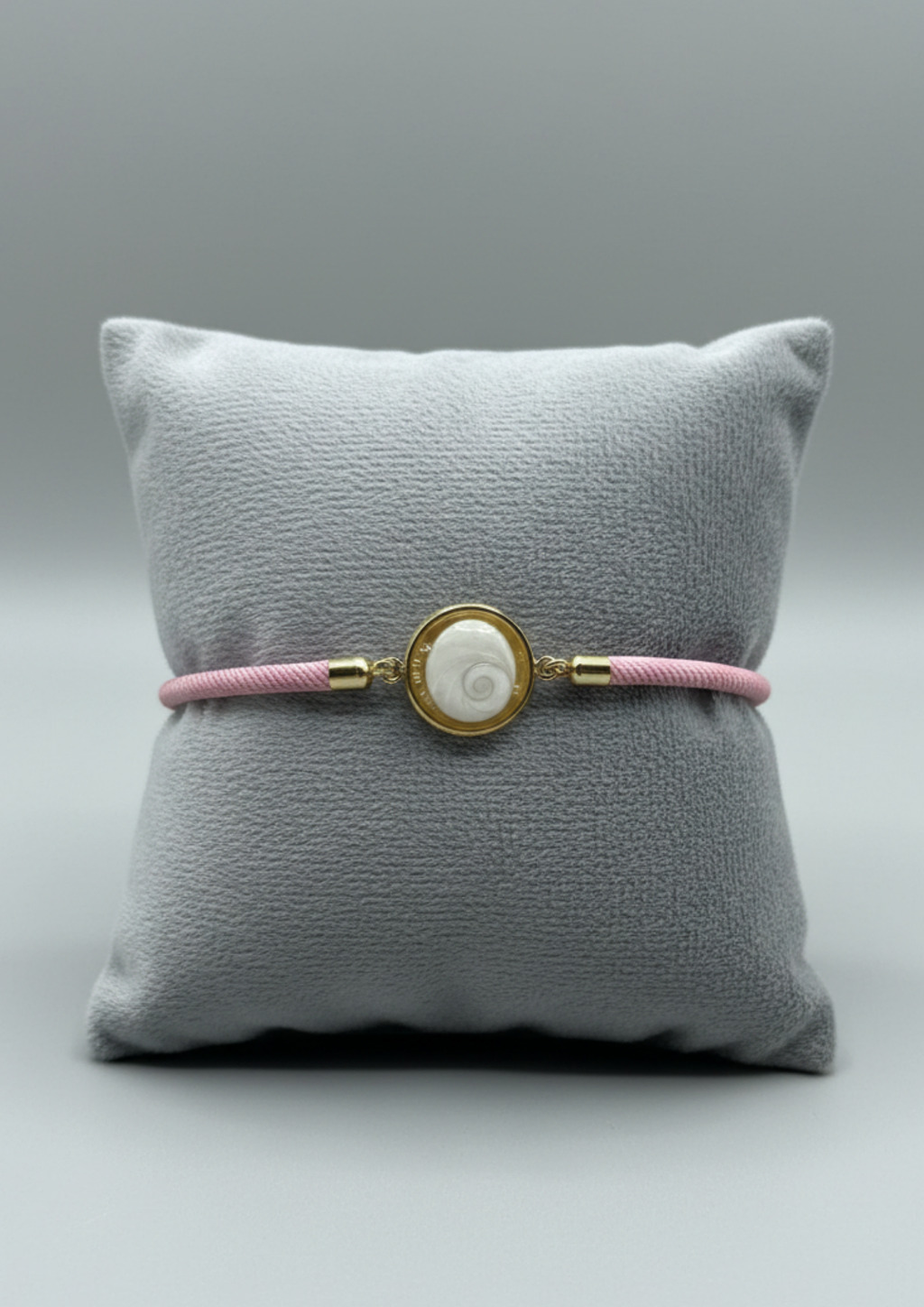 Bracelet femme sur coussin gris, cordon rose fin, médaillon rond doré serti d’un Œil de Sainte‑Lucie blanc, style chic et minimaliste.